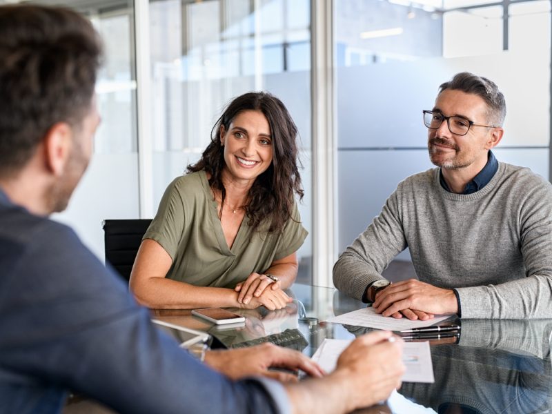 Smiling mature couple meeting with bank manager for investment. Beautiful mid adult woman with husband listening to businessman during meeting in conference room in modern office. Happy middle aged couple meeting loan advisor to buy a new home.